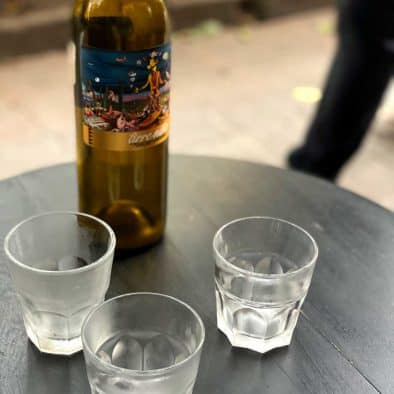 Bottle of wine with three glasses on a street table, vibrant street scene in background.