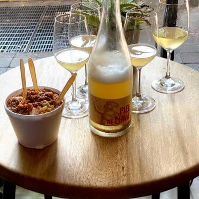 A bottle of white wine, three glasses, and a bowl of street food on a wooden table.